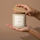 Hand holding a beige candle labeled 'rouge' with a wooden lid against a brown background