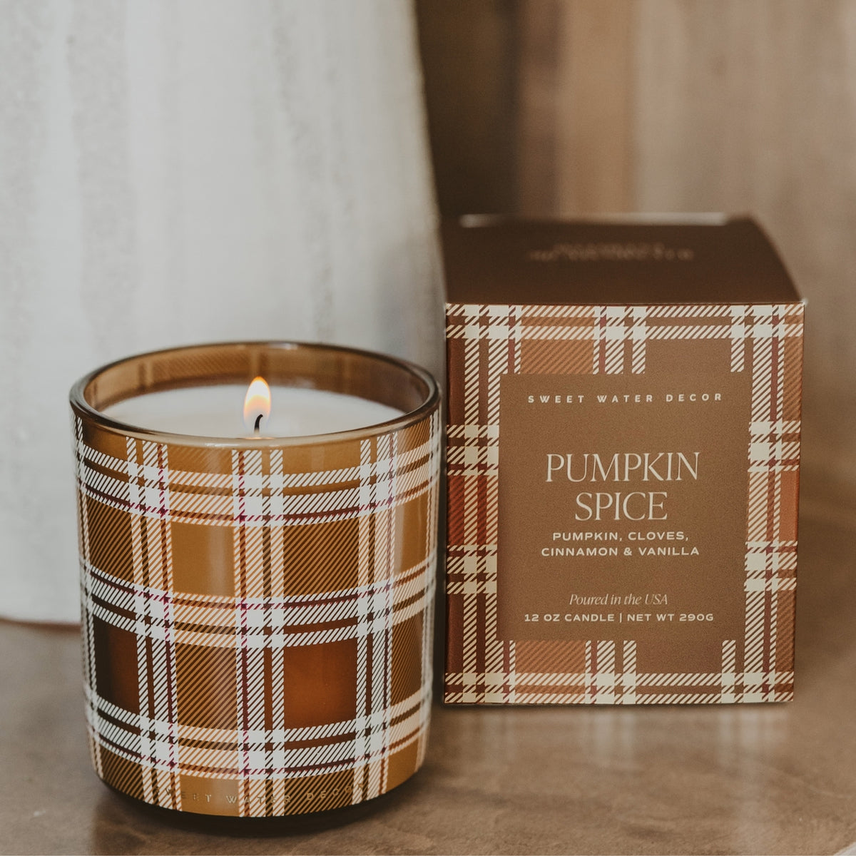 A lit candle in a brown plaid glass jar sits next to its matching retail box on a wooden surface. The box reads 'Sweet Water Decor' and 'Pumpkin Spice'.