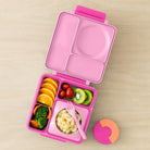 Pink bento box with compartments filled with food on a wooden surface, featuring OmieBox branding and certification logos.