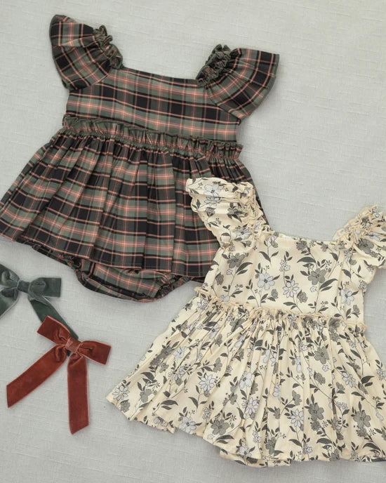 Two baby girl's off-the-shoulder dresses laid flat on a light gray fabric background. The top dress is dark brown and muted red plaid with ruffled shoulder straps and matching bloomers.