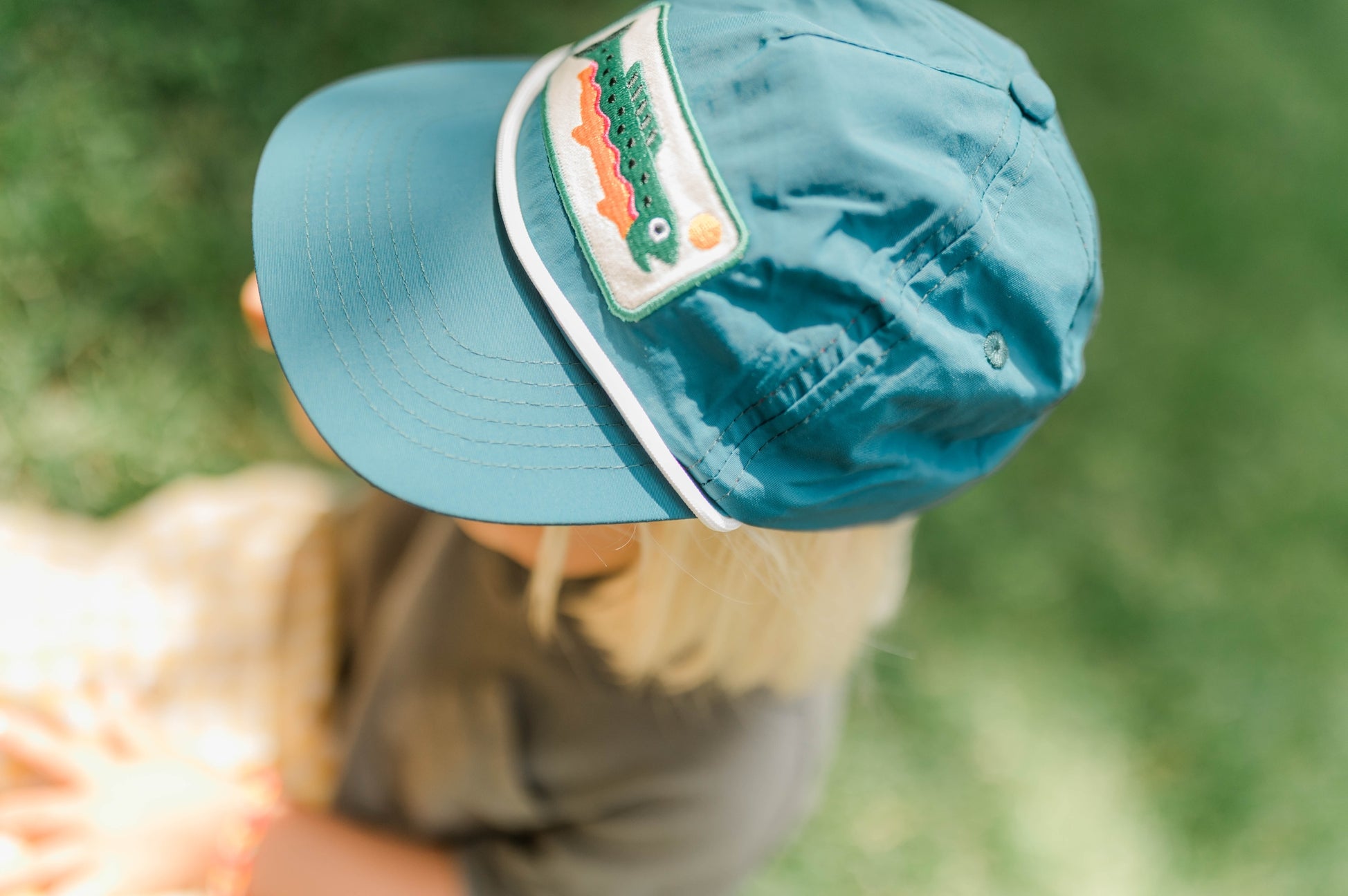 A close-up shot of a child wearing a teal blue baseball cap with a green alligator patch on the side, set against a soft-focus green outdoor background.