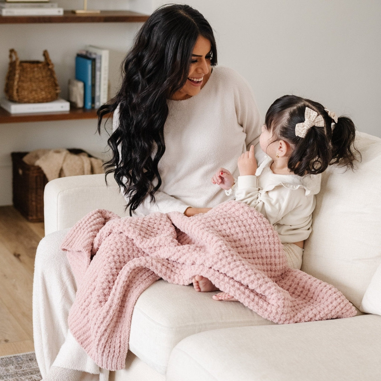 A woman and a young girl sit together on a cream sofa, wrapped in a dusty pink textured knit blanket. The woman wears a cream knit sweater, and the girl wears a cream top with a white bow in her ponytail.