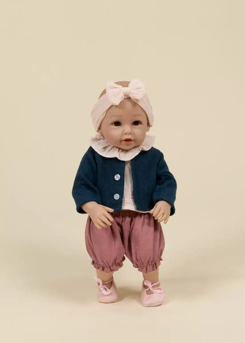 Baby doll wearing a navy blue cardigan, pink pants, and a matching headband on a beige background