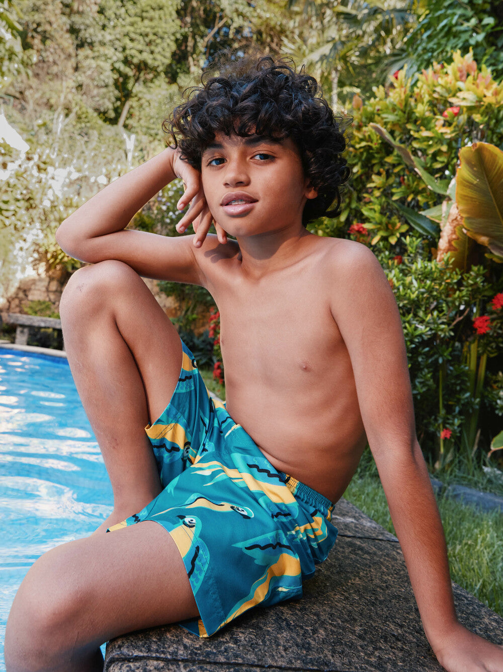 Young boy wearing blue swim trunks with yellow patterns, sitting by a pool.