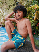 Young boy wearing blue swim trunks with yellow patterns, sitting by a pool.