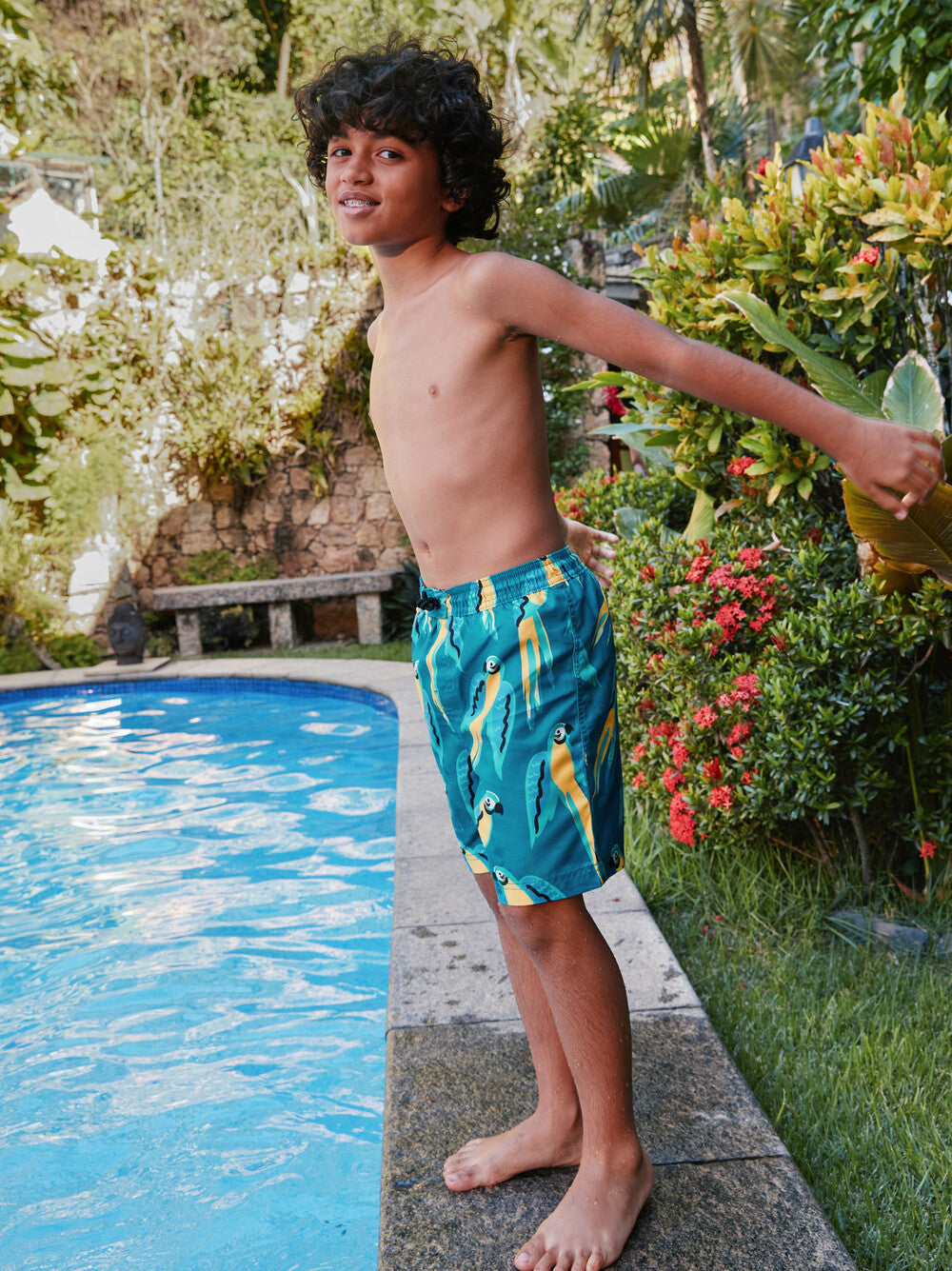 Child in swim shorts standing by a pool with greenery around