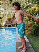 Child in swim shorts standing by a pool with greenery around