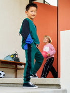 Child in green and navy tracksuit standing on steps with another child in pink shirt in the background.