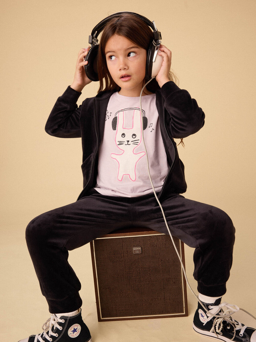 Child wearing headphones sitting on a speaker with a neutral background