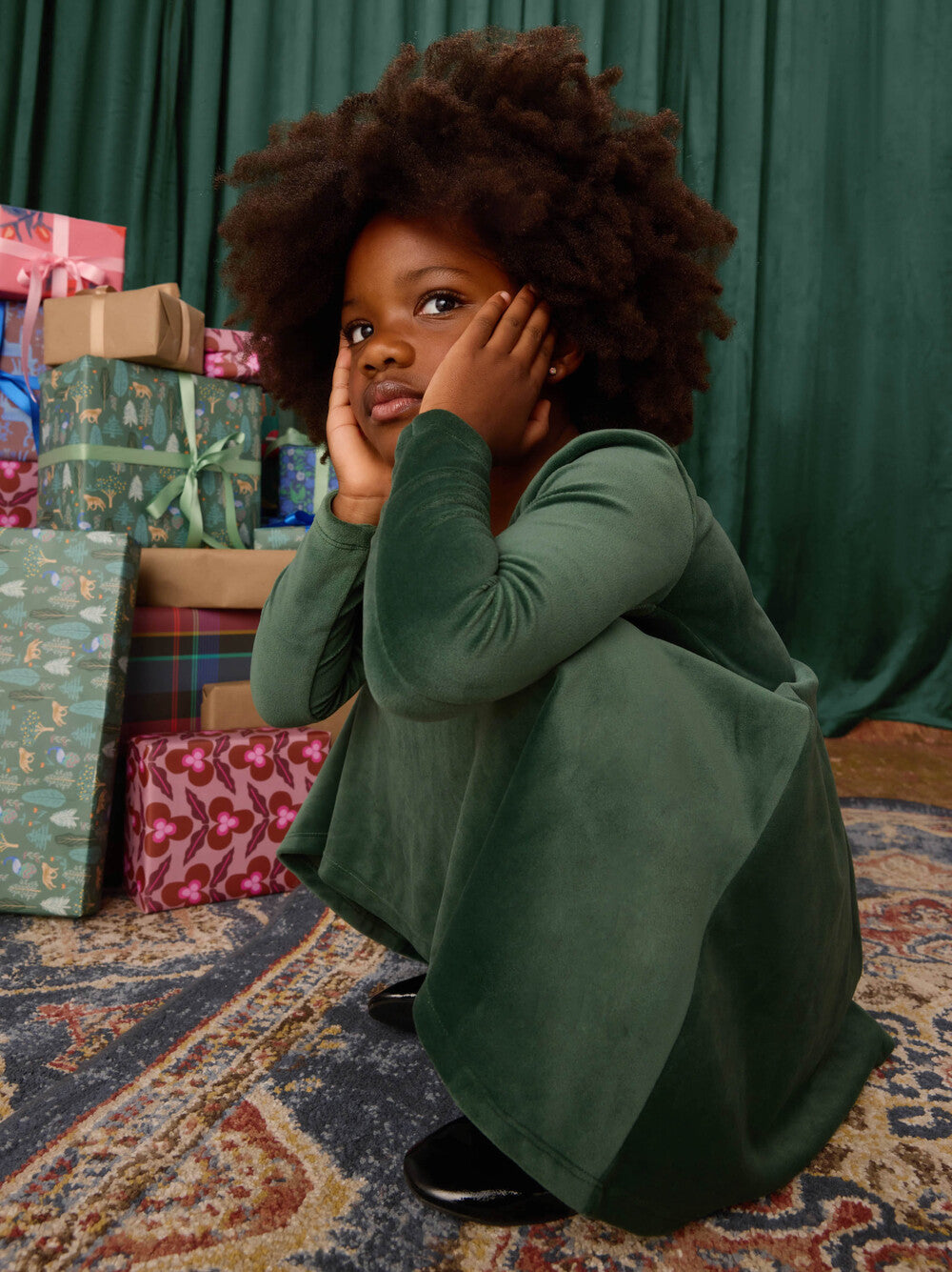 Child in a green outfit sitting on a patterned rug with gift boxes in the background