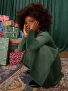 Child in a green outfit sitting on a patterned rug with gift boxes in the background