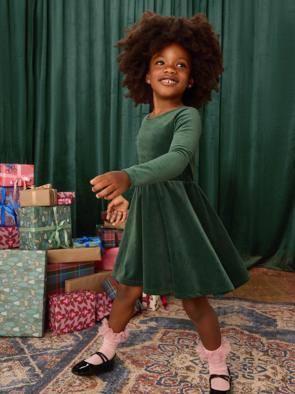 Young girl in a green dress standing in front of Christmas presents with a green curtain background