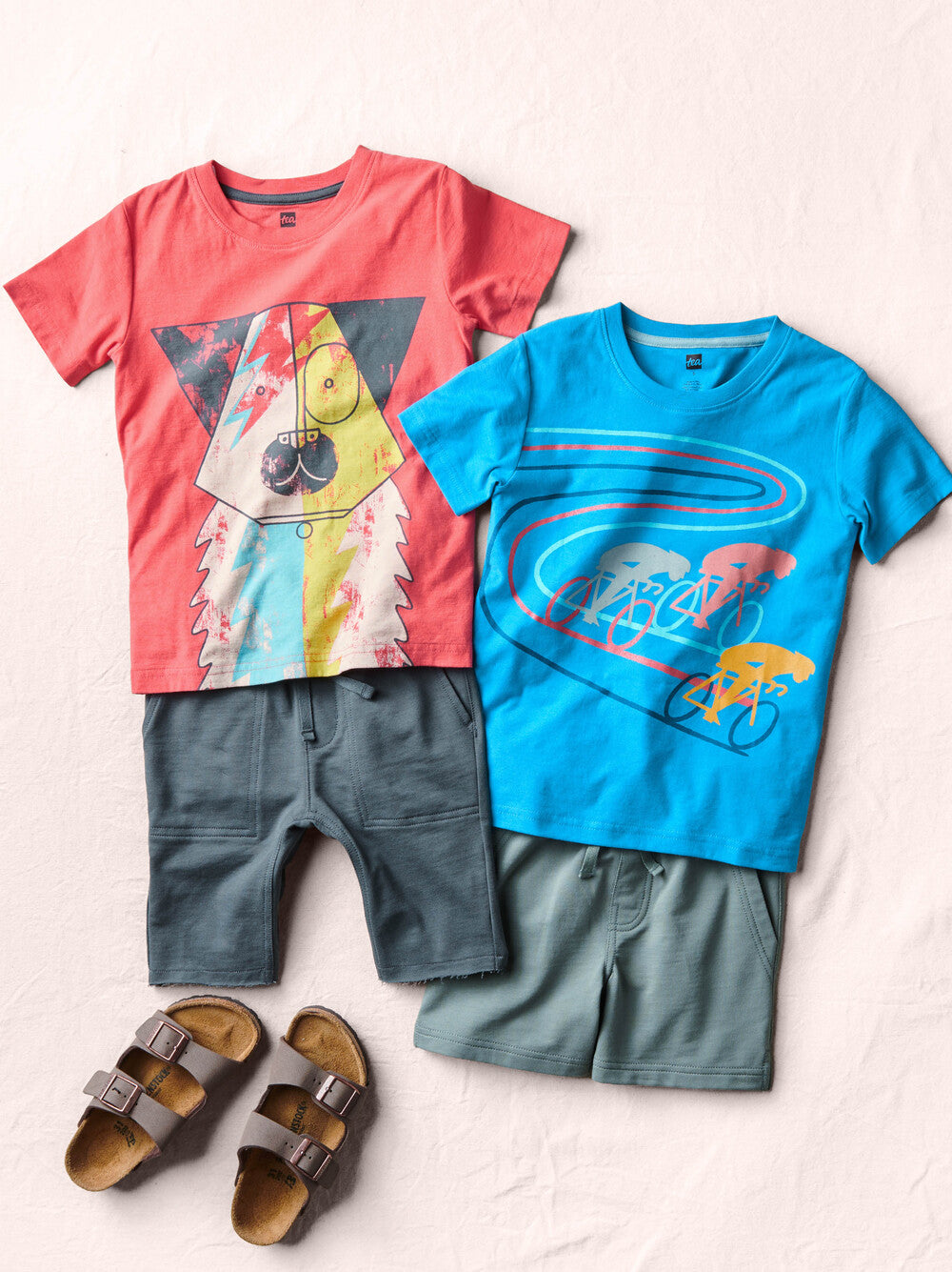 Two children's t-shirts with graphic designs and shorts, paired with sandals on a light background.