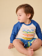 Child wearing a blue and white long-sleeve shirt with colorful design, sitting on a beige background