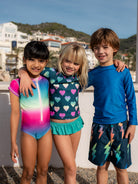 Three children in colorful swimwear standing together outdoors with a scenic background.