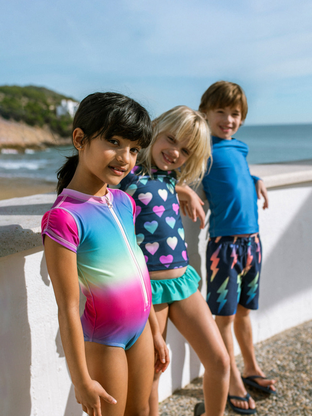 Three children in colorful swimsuits standing by a beach.