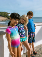 Three children in colorful swimsuits standing by a beach.