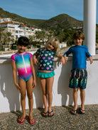 Three children in swimsuits standing against a white wall with a scenic background.