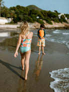 Two children on a beach with one walking towards the water and the other holding an orange float.
