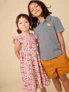 Two children, a girl in a floral dress and a boy in a striped shirt and shorts, standing against a beige background.