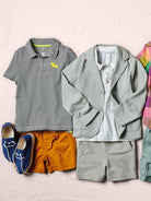 Children's clothing including a striped polo shirt, jacket, shorts, and shoes on a light pink background.