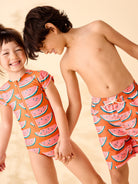 Two children wearing watermelon-patterned swimsuits on a beige background