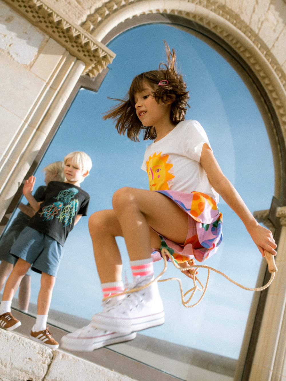 Two children playing on a rope in front of a large window with a clear blue sky.