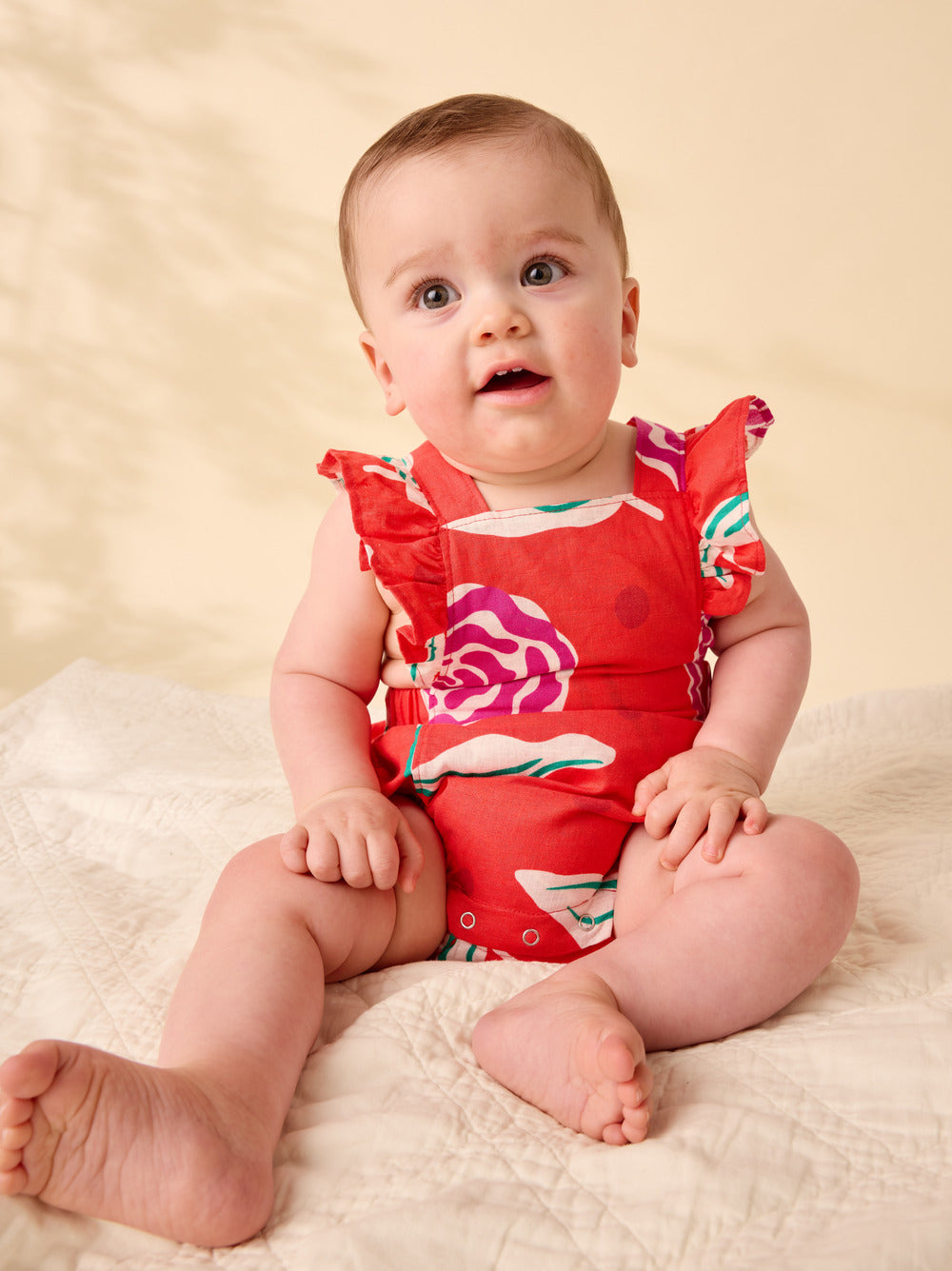 Baby wearing a red floral swimsuit sitting on a beige blanket.