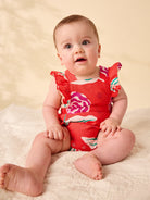Baby wearing a red floral swimsuit sitting on a beige blanket.