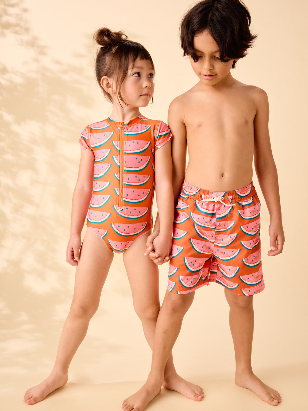 Two children wearing watermelon-patterned swimsuits on a beige background