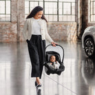 Woman pushing a baby in a car seat carrier in an indoor setting with a car visible.
