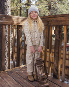 A young girl stands on a wooden deck wearing a beige and brown animal-print hooded one-piece outfit, a gray knit beanie, and tan boots, holding a vintage-style lantern. Trees and a wooden fence are visible in the background.