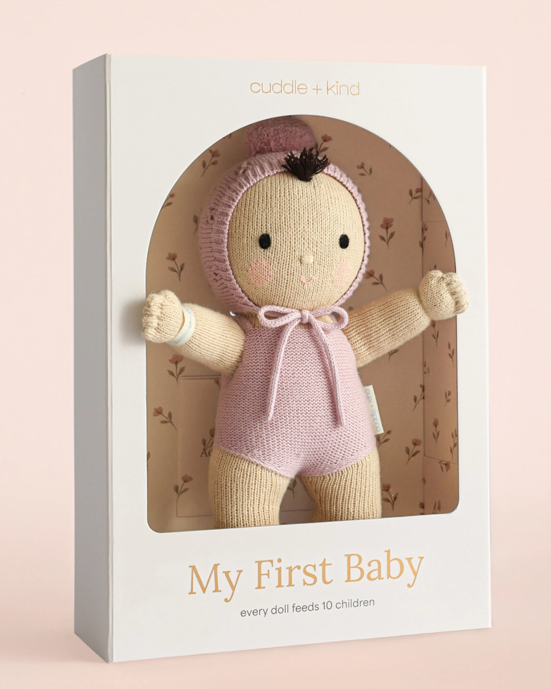 A knitted baby doll in a pink outfit and bonnet displayed inside a white gift box with a window. The box has the brand name 'cuddle + kind' at the top and the title 'My First Baby' at the bottom, set against a soft pink background.