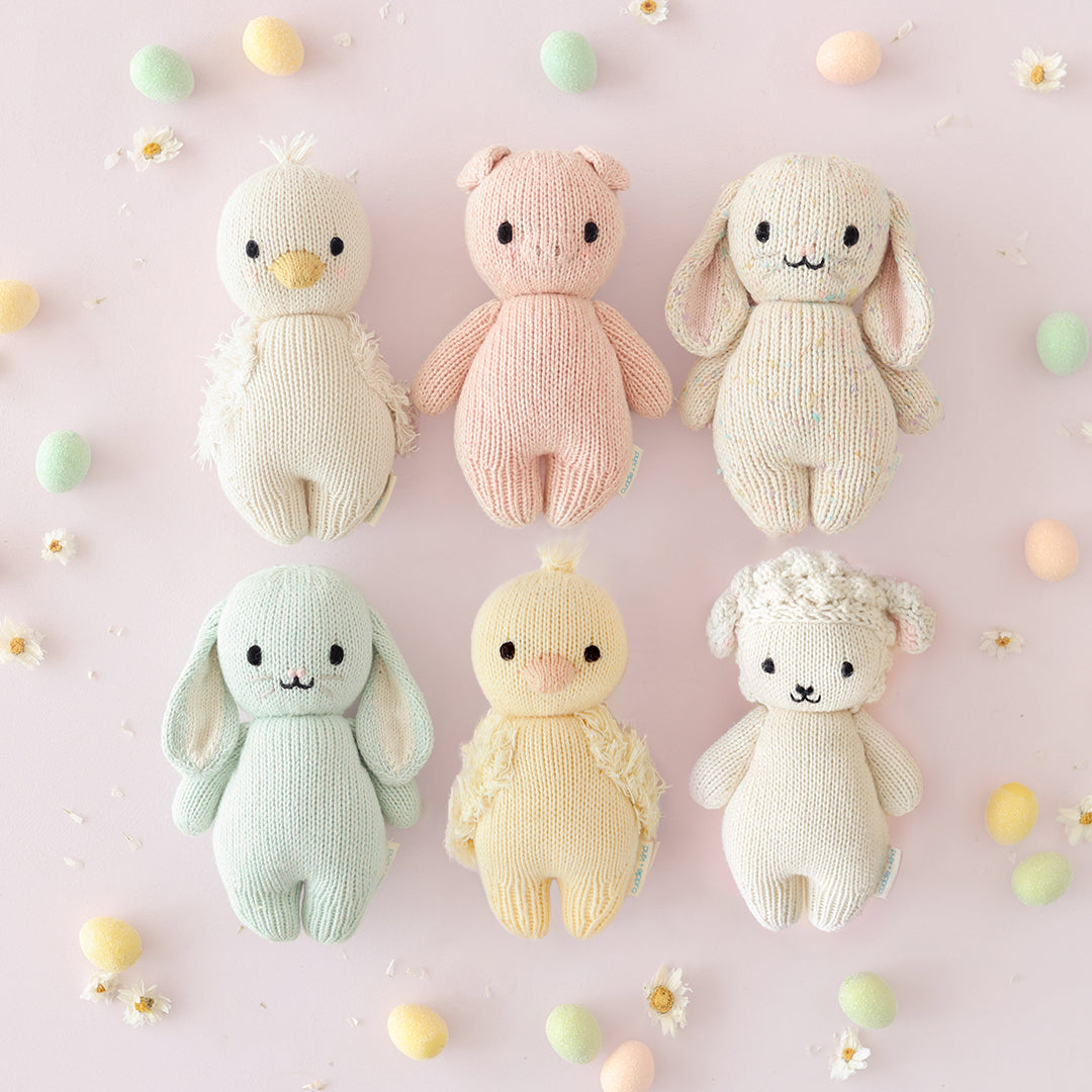 Six knitted toys in pastel colors on a light pink background with small colorful balls and flowers.