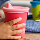 Hand holding a pink sippy cup on a wooden surface with a blurred background