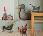 A miniature green wooden Maileg toy mouse sleigh on a wooden floor, with a larger plush toy duck, gnome dolls, a small suitcase, and holiday decor arranged in a rustic room setting.