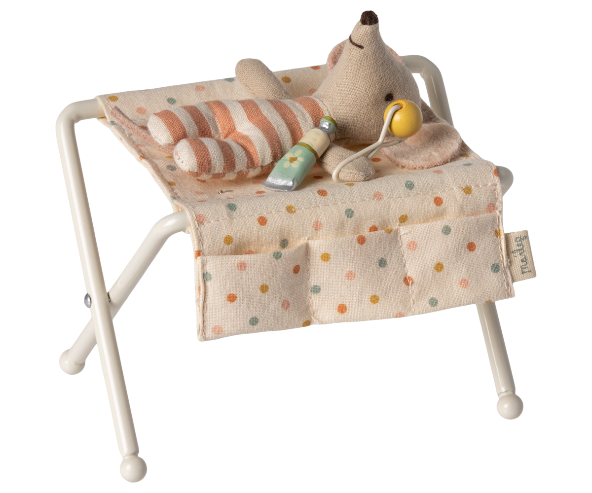 Maileg off-white miniature nursery changing table for baby mice, featuring metal folding legs, a polka-dot fabric changing pad, side pockets, a tiny rattle, and a baby lotion bottle.