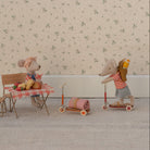 Two toy mice with a table and chairs, one on a scooter with a backpack, against a floral wallpaper background.