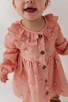 A close-up of a toddler wearing a dusty pink long-sleeve dress with a ruffled collar, button front, and a small mushroom print, against a plain light background.