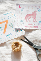 Two decorative printed paper cards with scalloped edges laid on a white fabric surface, alongside a small ball of twine and scissors. One card shows a pink wolf illustration with the text 'LOBO MEXICANO' and the letter 'L', while the other shows a large orange numeral '7' and the word 'SIETE'.