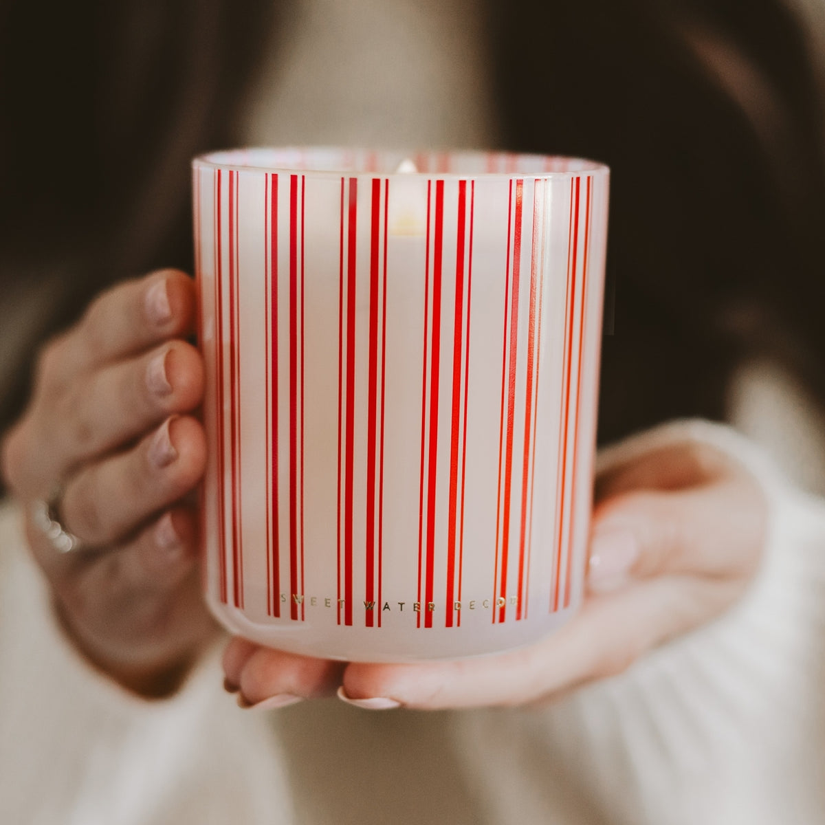 A close-up of a person holding a frosted glass candle jar with red and white vertical stripes. The person is wearing a white knit sweater, and the candle is lit with a warm glow.