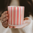 A close-up of a person holding a frosted glass candle jar with red and white vertical stripes. The person is wearing a white knit sweater, and the candle is lit with a warm glow.