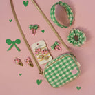 A flat-lay of a green and white gingham crossbody bag with a gold strap, accompanied by matching hair accessories including a headband, scrunchie, and bow clips, all arranged on a pink background with decorative green hearts.