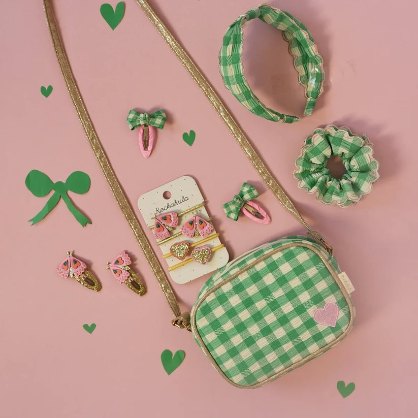 A flat-lay of a green and white gingham crossbody bag with a gold strap, accompanied by matching hair accessories including a headband, scrunchie, and bow clips, all arranged on a pink background with decorative green hearts.