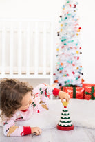 A young child wearing pink Christmas pajamas lies on a white rug, stacking a small wooden Christmas tree toy. A decorated white Christmas tree and red wrapped gifts are visible in the softly blurred background.
