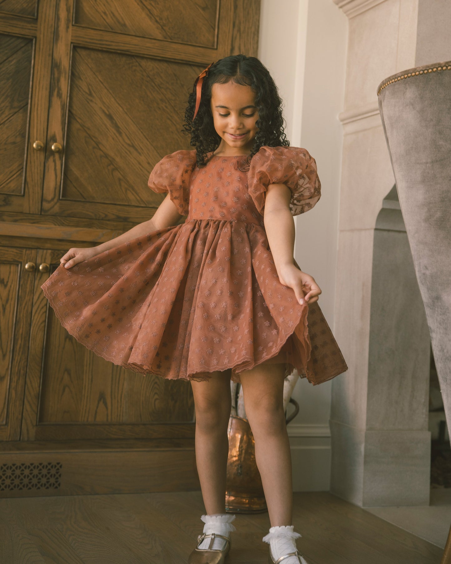 A young girl with curly dark hair wearing a rust-orange polka dot dress with puff sleeves, twirling in a room with a wooden cabinet and a gray velvet chair.