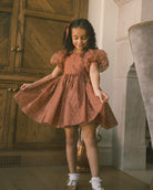 A young girl with curly dark hair wearing a rust-orange polka dot dress with puff sleeves, twirling in a room with a wooden cabinet and a gray velvet chair.