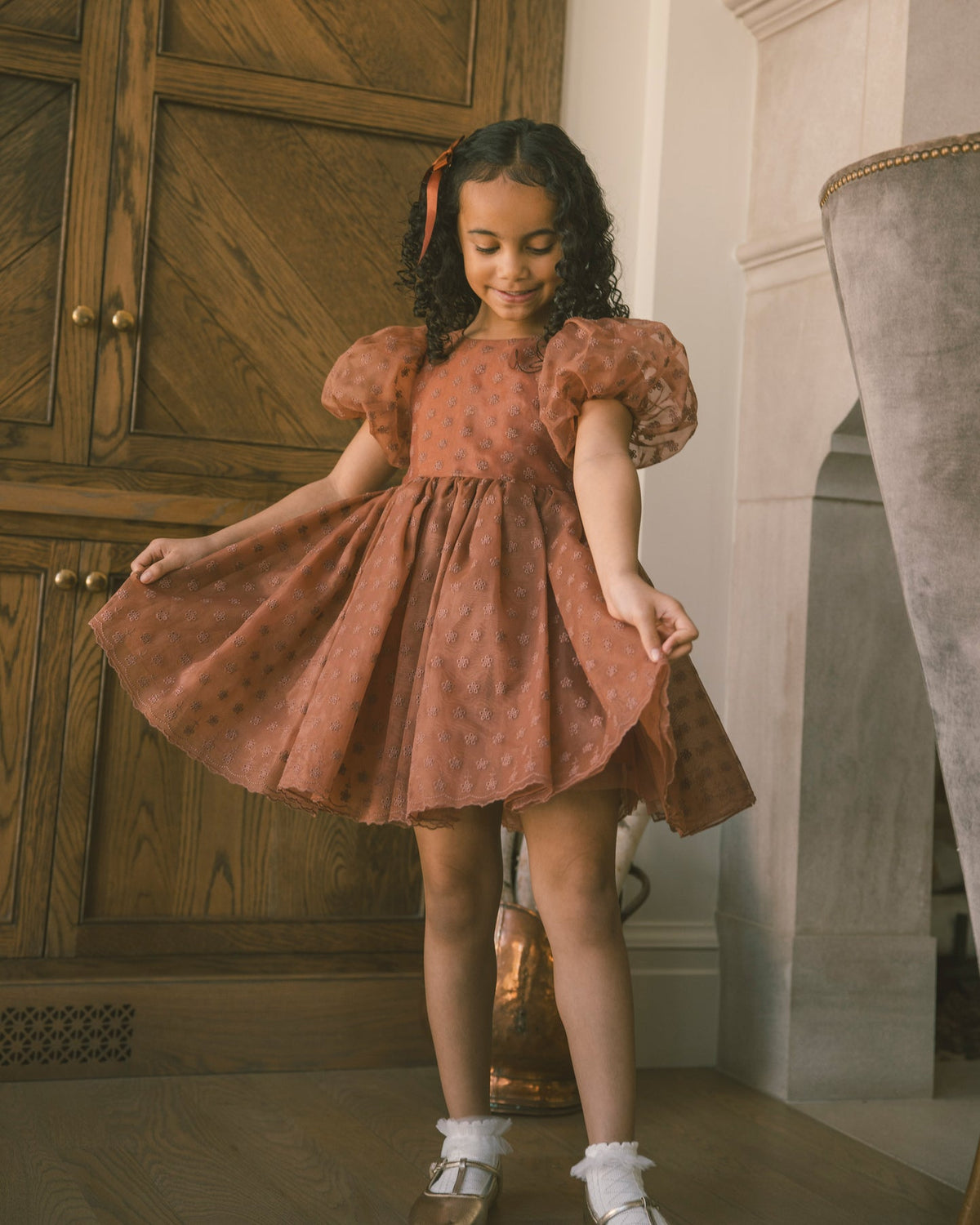 A young girl with curly dark hair wearing a rust-orange polka dot dress with puff sleeves, twirling in a room with a wooden cabinet and a gray velvet chair.