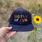 Navy blue cap with colorful text 'BIG FAN OF FUN' held in front of sunflowers.