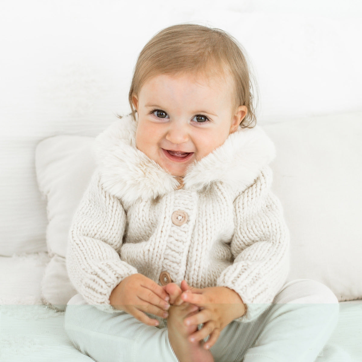 Smiling baby wearing a white knit cardigan on a light background